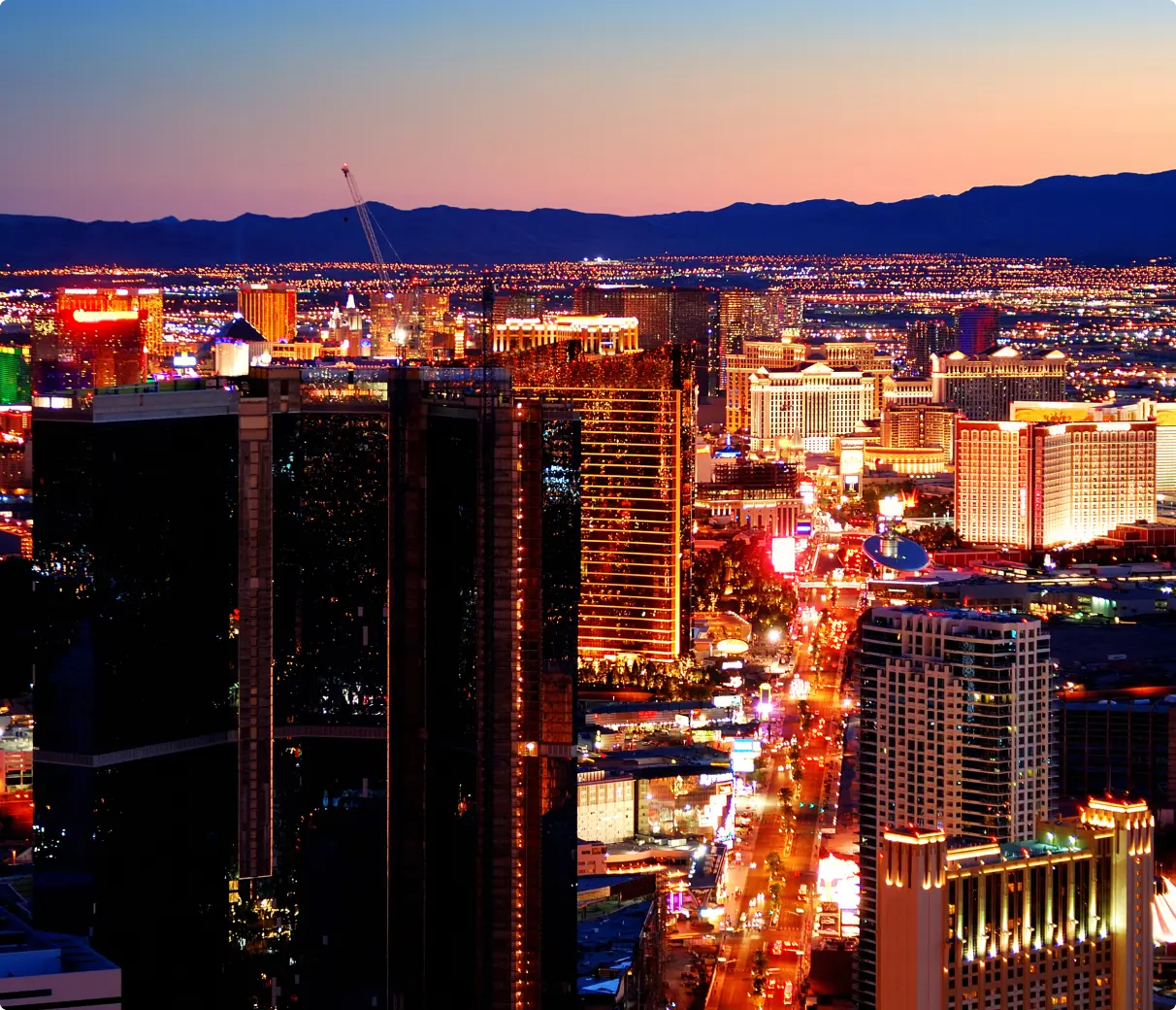Aerial view of Las Vegas at dusk, with the Strip brightly illuminated. Skyscrapers and hotels glow against a darkening sky, creating a vibrant, lively atmosphere.