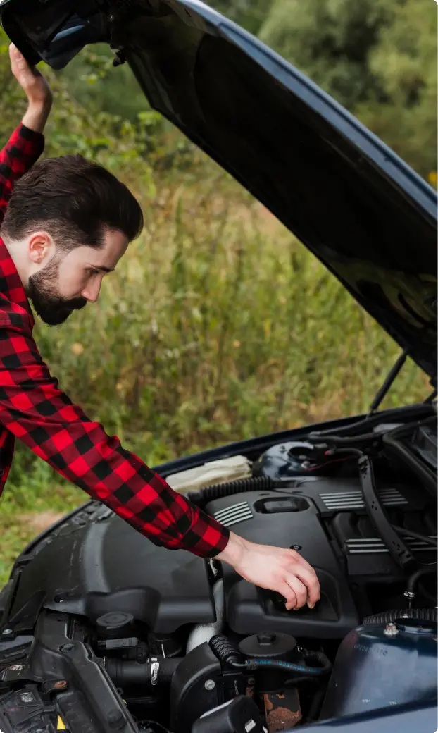 A man in a red plaid shirt inspects a car engine with the hood open in a rural setting. He appears focused, examining the engine components.