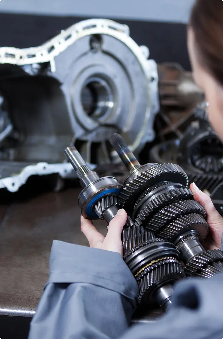 Person in a workshop holding a complex set of metal gears and shafts. A transmission case is visible in the background. The scene is focused and technical.