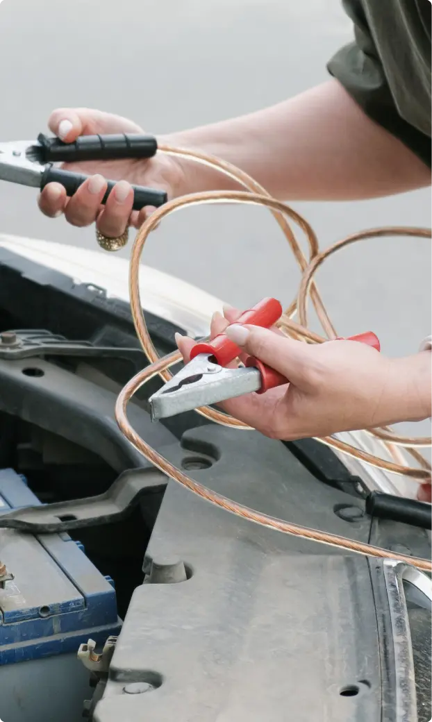 Close-up of hands holding jumper cables, connecting to a car battery. The scene conveys focus and readiness, suggesting a roadside assistance situation.