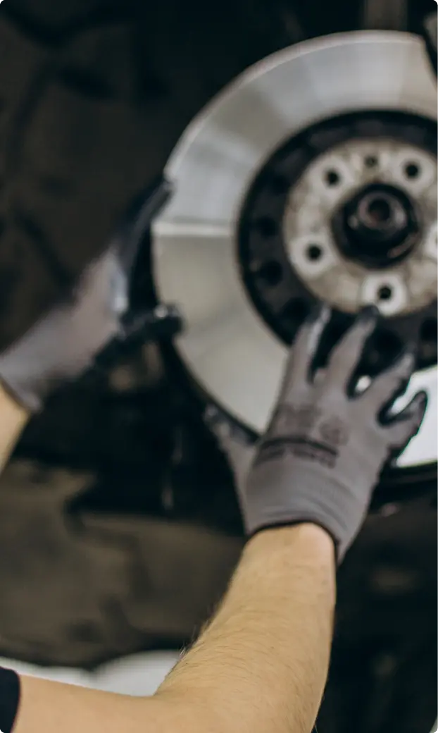 Close-up of a mechanic's arms wearing gray gloves, installing a shiny, silver brake disc onto a vehicle. The scene conveys precision and focus.