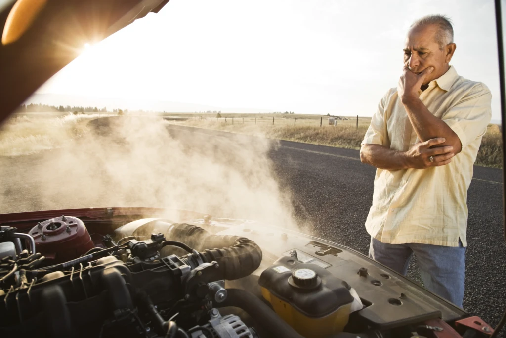 Section 1An older man stands on a rural road, thoughtfully observing steam rising from his car's engine. The sun sets in the background, creating a concerned yet contemplative mood.