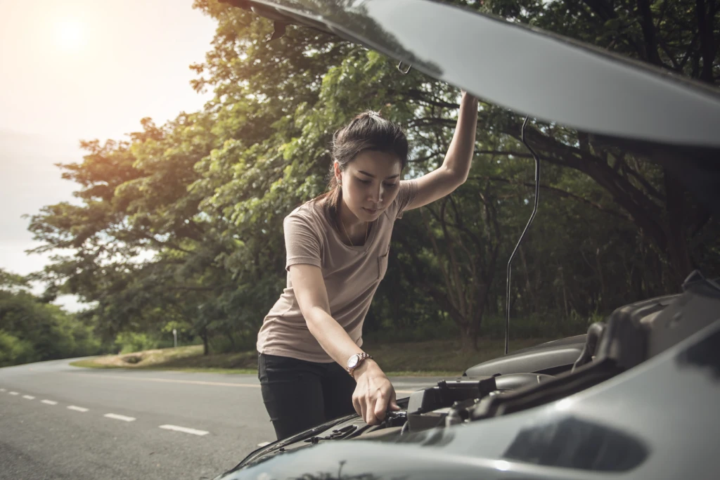 A woman leans over the open hood of a car on a rural road, inspecting the engine. Sunlight filters through nearby trees, creating a warm, thoughtful scene.